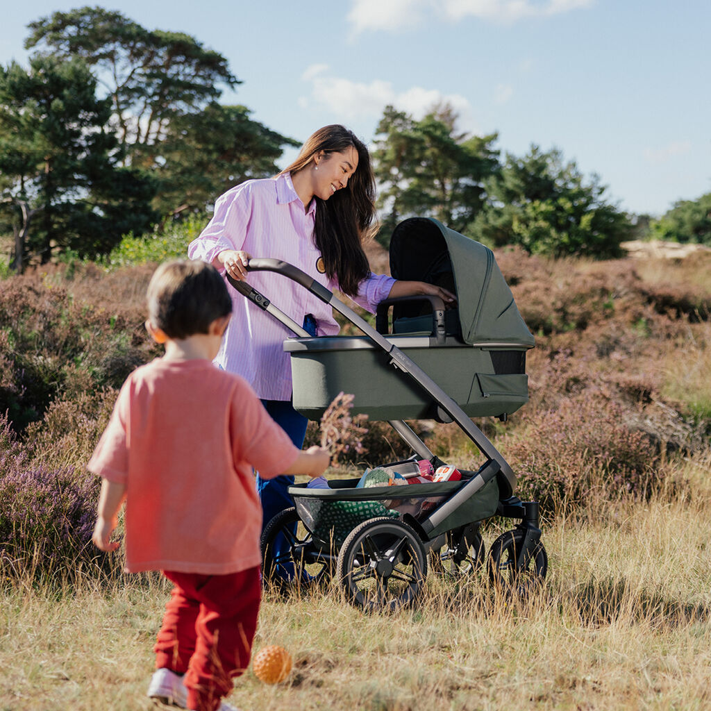 Parent pushing a Joolz stroller with baby carrycot through the countryside, with a child walking alongside.