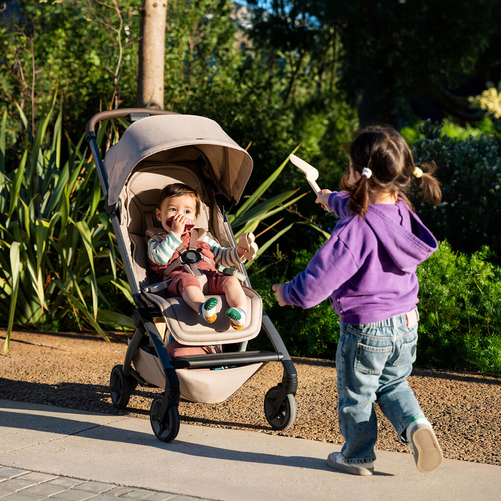 Child sitting in a Joolz Aer2 stroller while another child plays alongside outdoors in a garden.